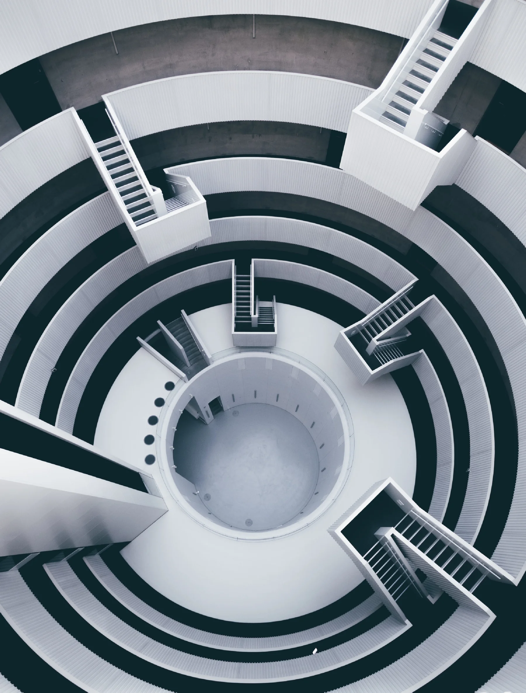 Top-down view of a circular, multi-level concrete structure with staircases radiating from a central void, echoing the labyrinth concept